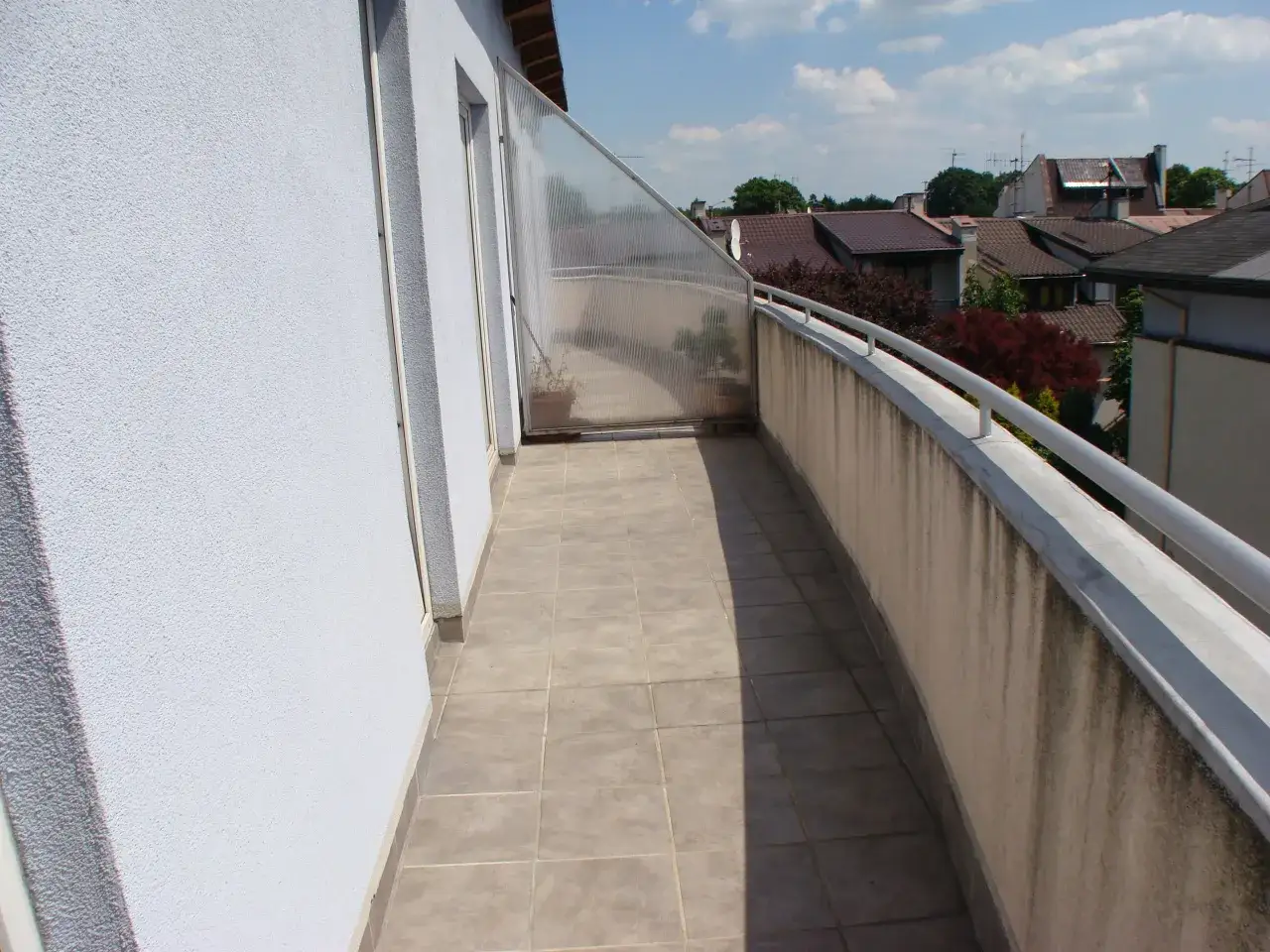 Długi balkon w bloku z wielkiej płyty, wyłożony płytkami, z przeszkloną ścianką i widokiem na dachy domów.