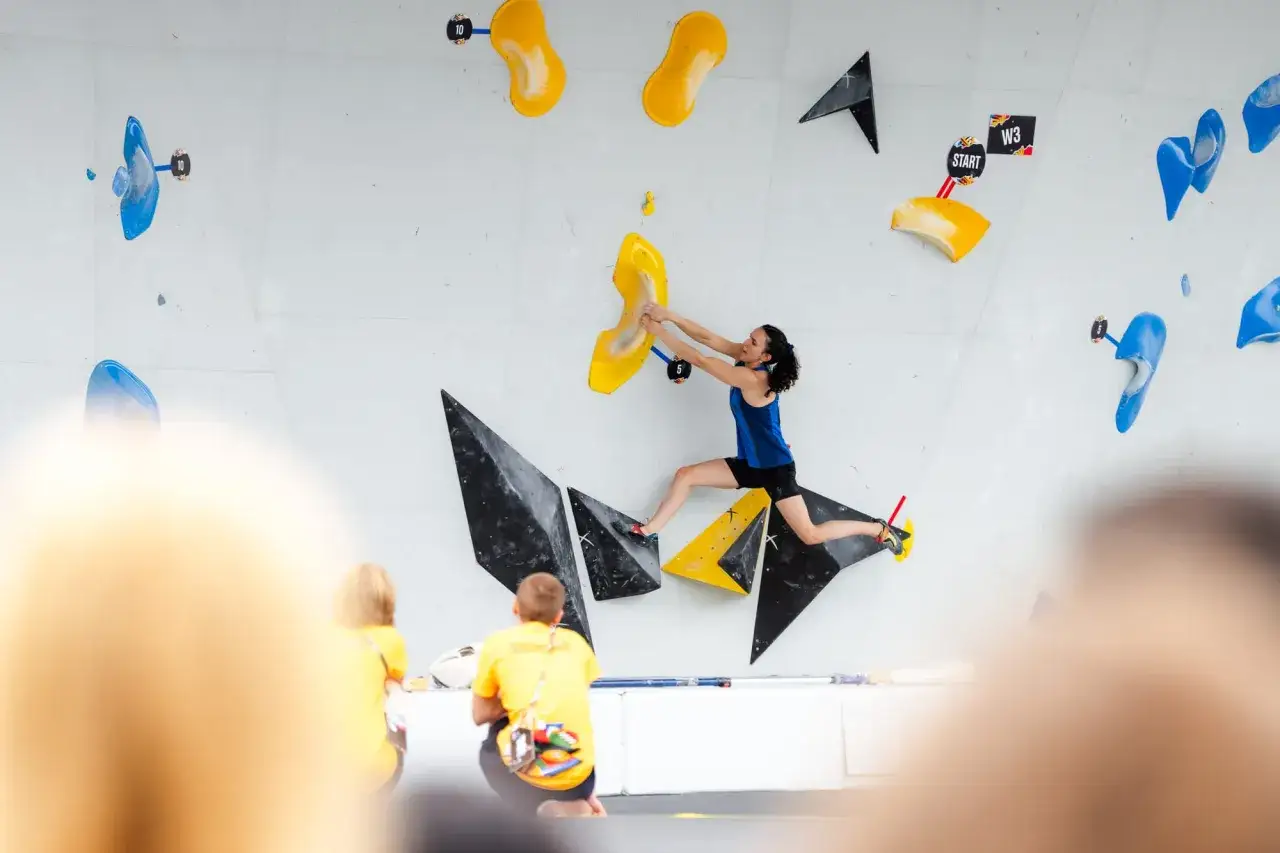 Kobieta w skoku na ścianie wspinaczkowej, demonstrująca dynamiczne rodzaje wspinaczki. Widzowie w żółtych koszulkach obserwują zmagania.