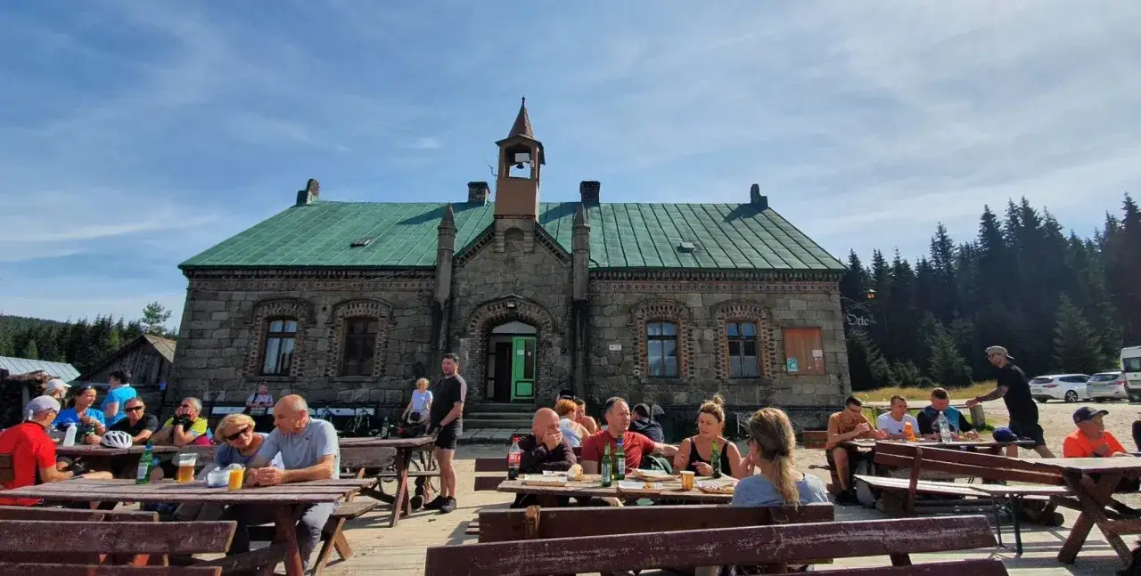 Ludzie odpoczywają przy stołach przed kamiennym budynkiem stacji turystycznej Orle.