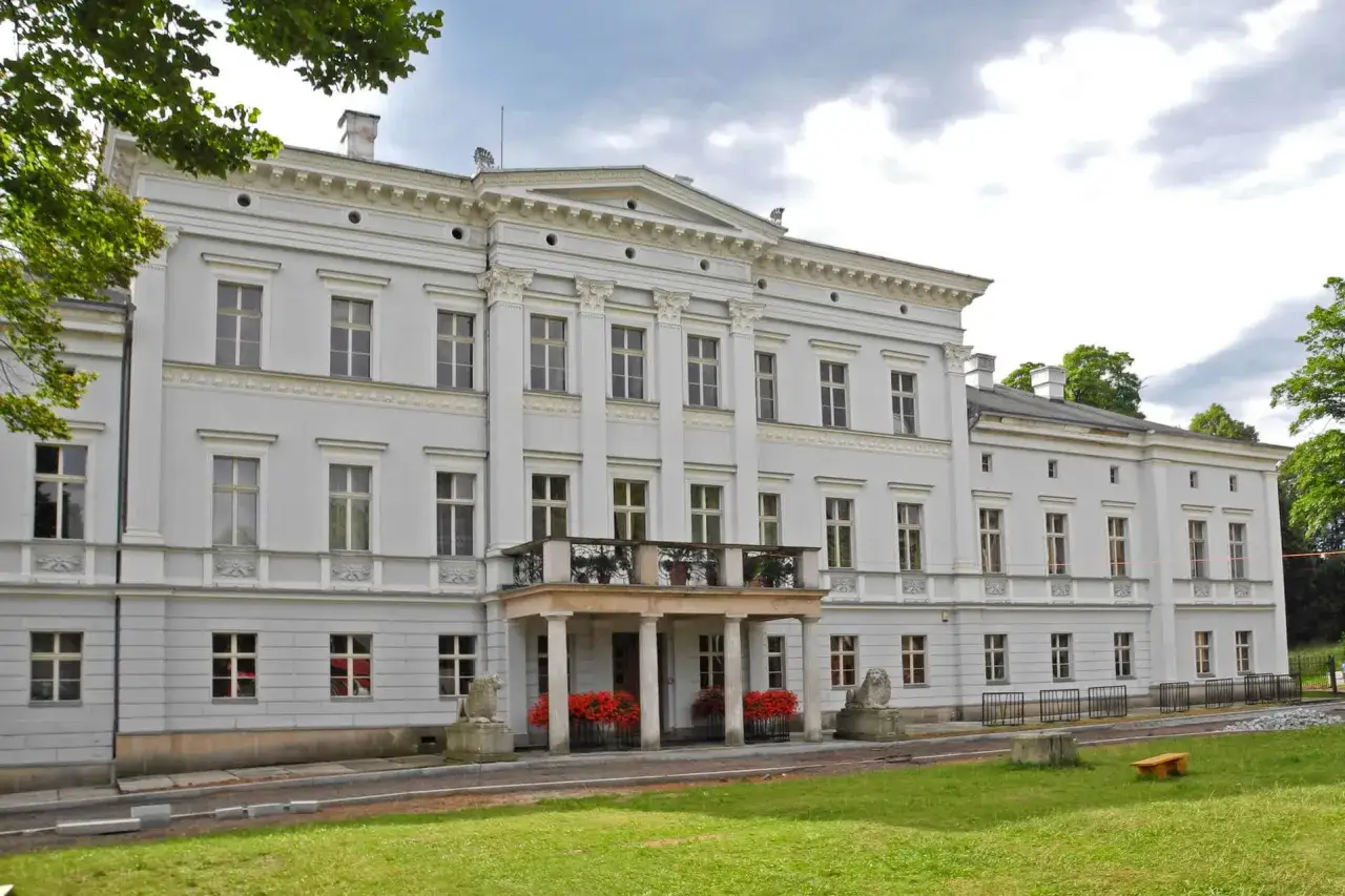 Elegancki pałac Jedlinka, z kolumnowym portykiem i lwami u wejścia, otoczony zielenią.