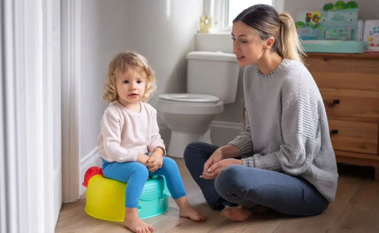 Mama rozmawia z dzieckiem siedzącym na kolorowym nocniku. To scena pokazująca, jak przekonać dziecko do nocnika.