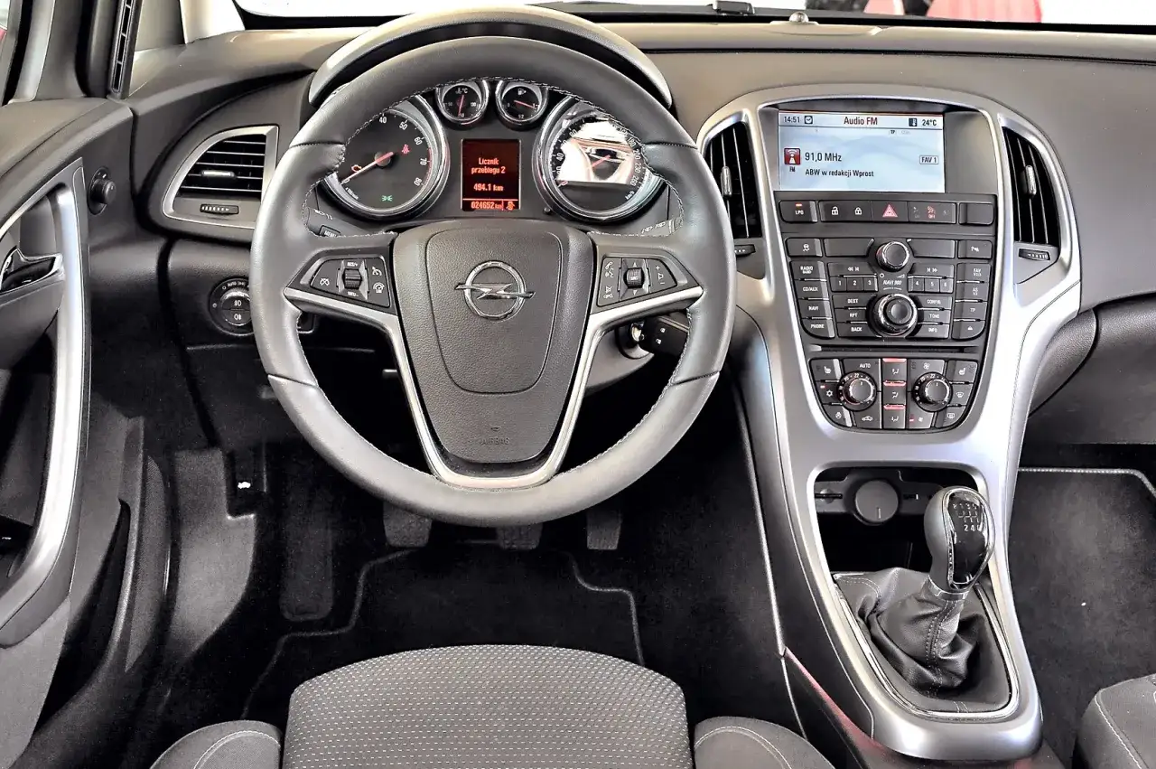 Wnętrze Opla Astry H z widocznym przyciskiem sport, kierownicą i panelem multimedialnym.