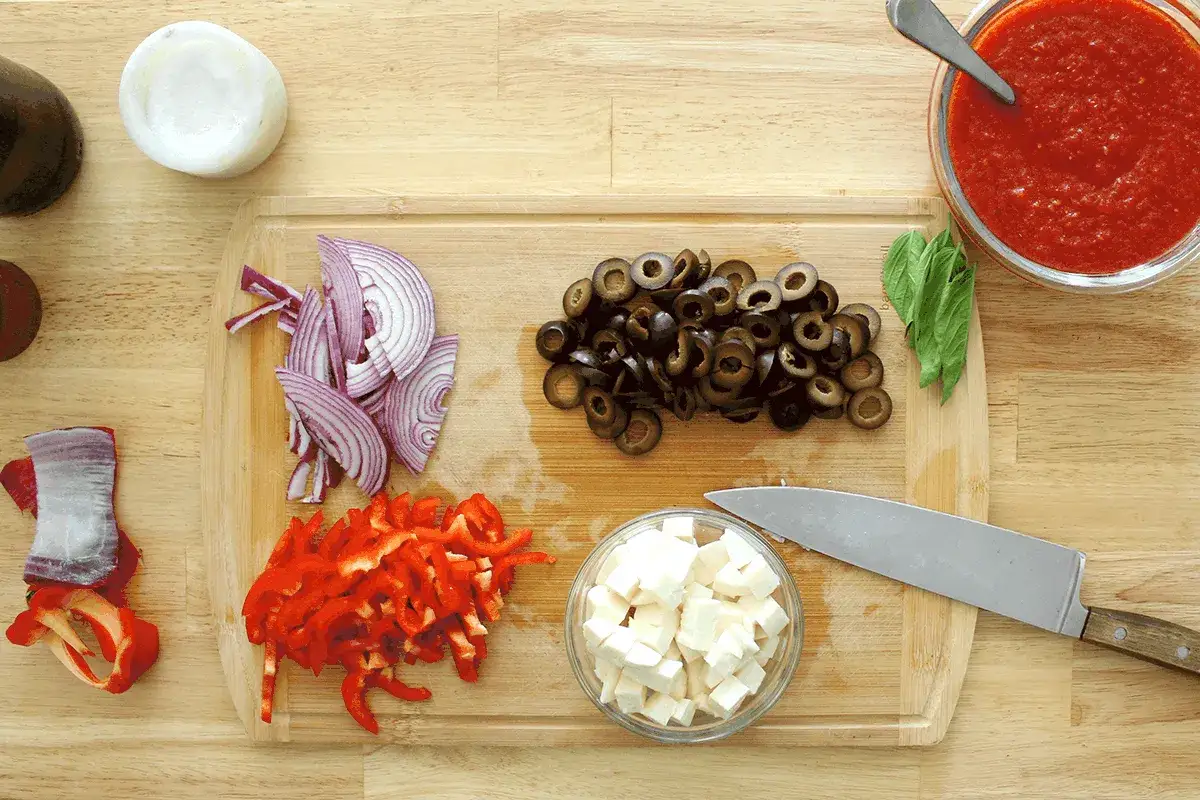 Pokrojone dodatki do pizzy: cebula, papryka, oliwki, ser. Obok sos pomidorowy i bazylia.