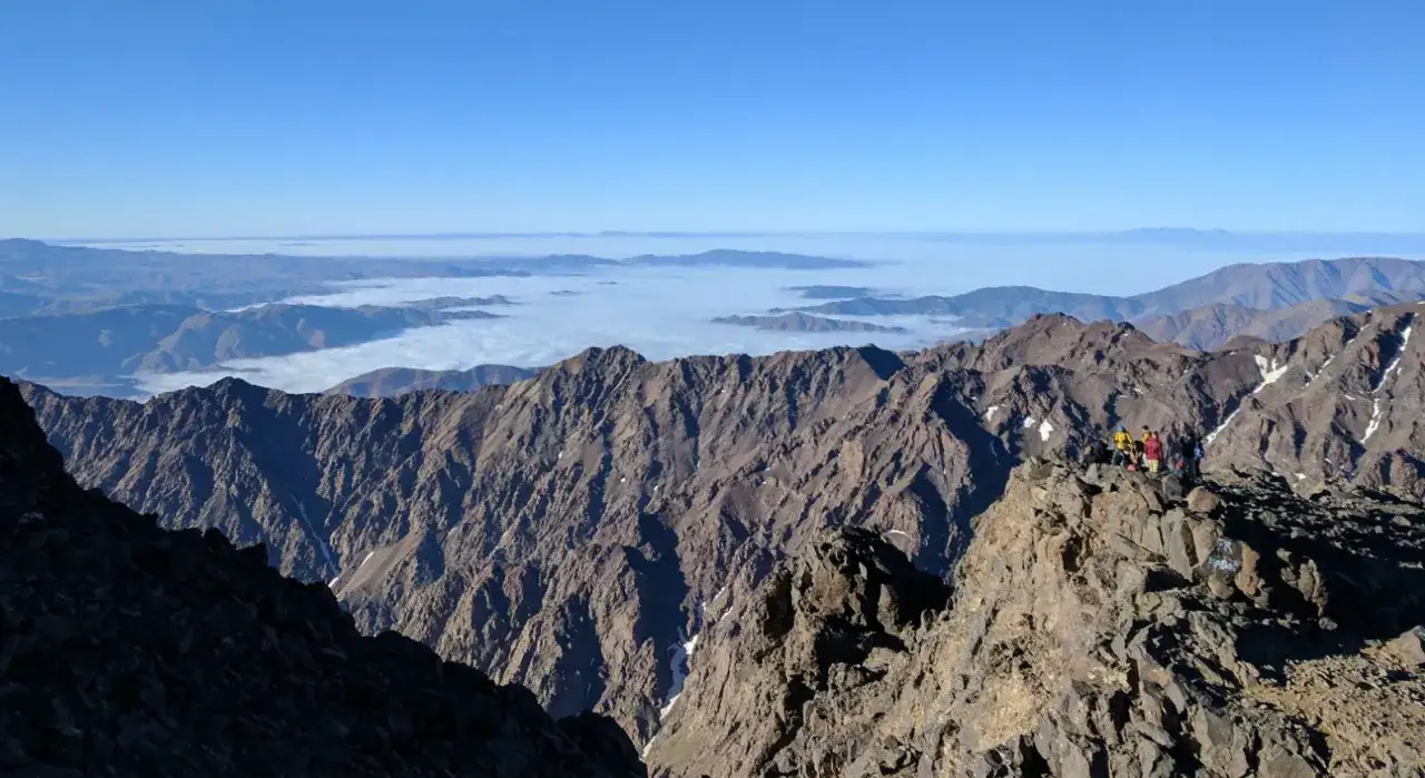 Grupa turyst&oacute;w na szczycie Jebel Toubkal, z widokiem na morze chmur i g&oacute;ry w oddali.