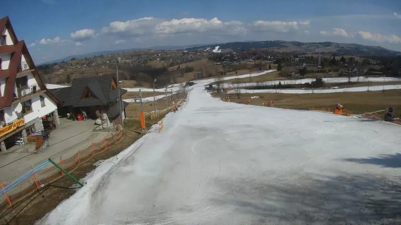 Widok z bukowina ski kamery na łagodny stok narciarski, z budynkami i górami w tle.