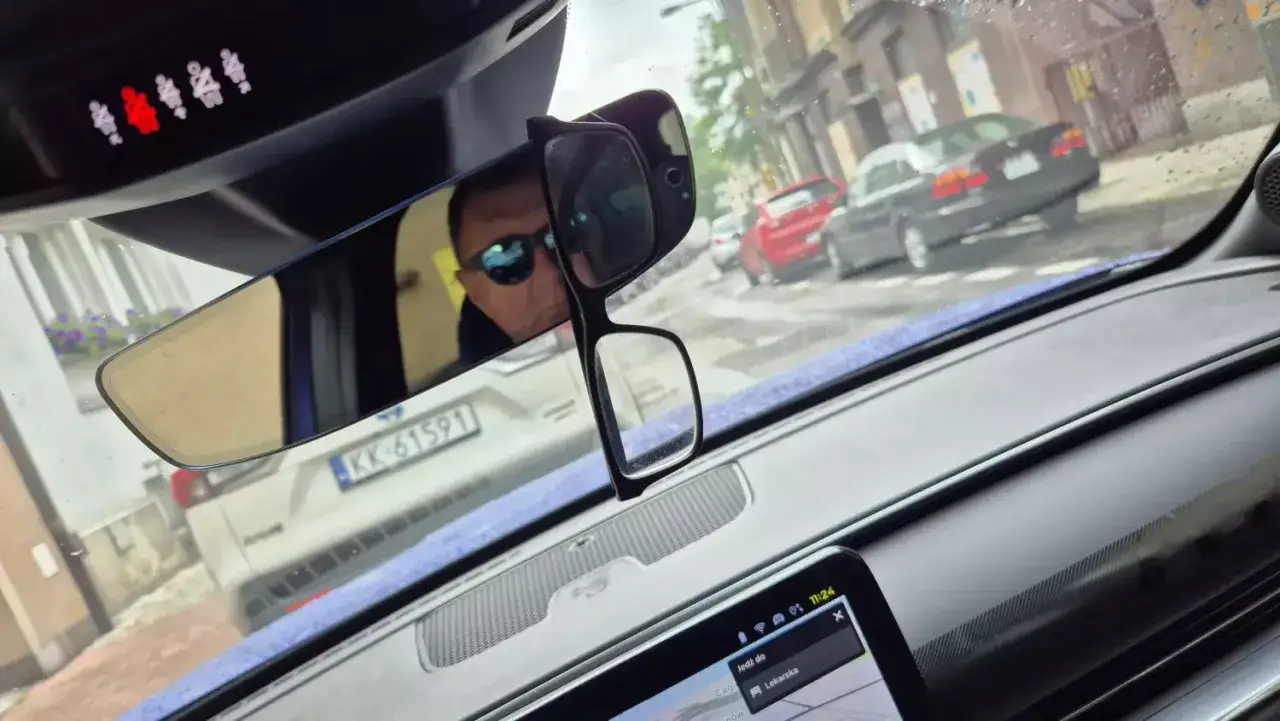 Okulary wiszą na lusterku wstecznym, zasłaniając widok. Kierowca w okularach przeciwsłonecznych prowadzi auto.