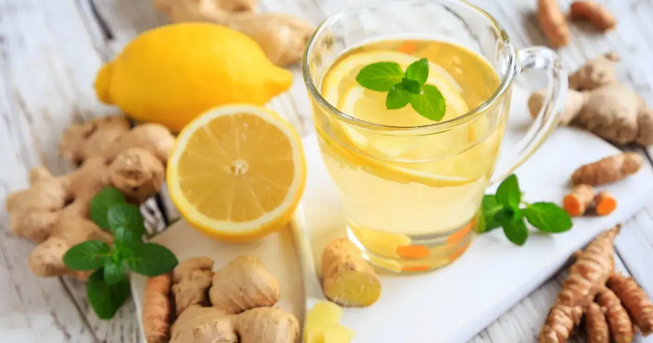 Jak zrobić herbatę z imbirem? Na stole leżą cytryny, imbir, mięta i kurkuma, a obok szklanka z napojem.