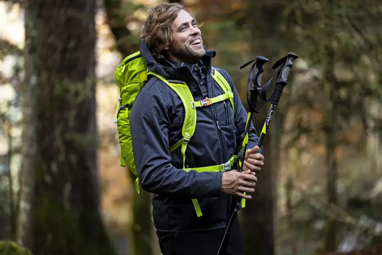 Szczęśliwy turysta w lesie, trzymający kije trekkingowe. Ma na sobie ciemny softshell z jaskrawozielonymi elementami plecaka i szelek.