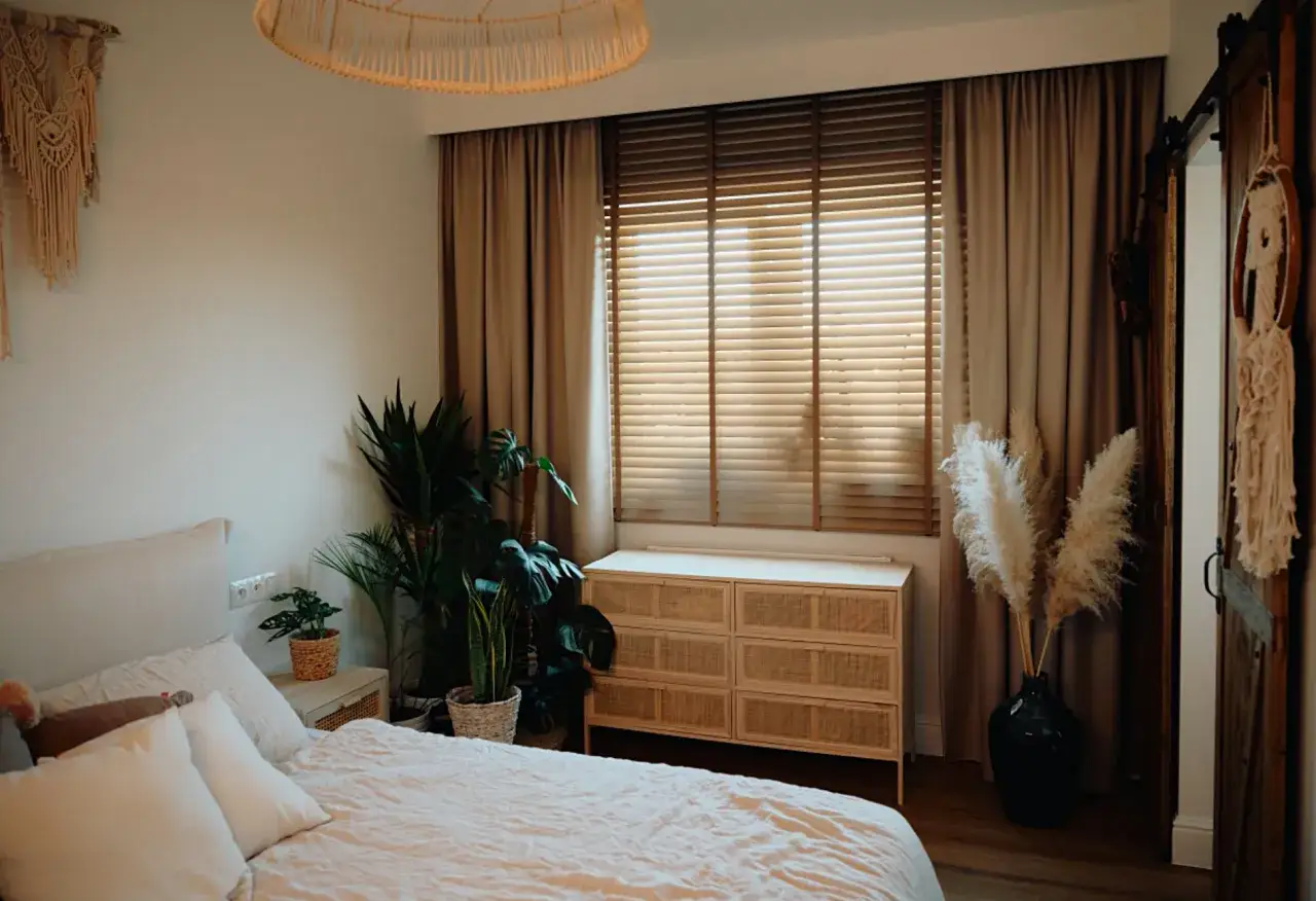 Sypialnia z żaluzjami drewnianymi i zasłonami w odcieniach beżu. Wnętrze zdobią rośliny i makrama.