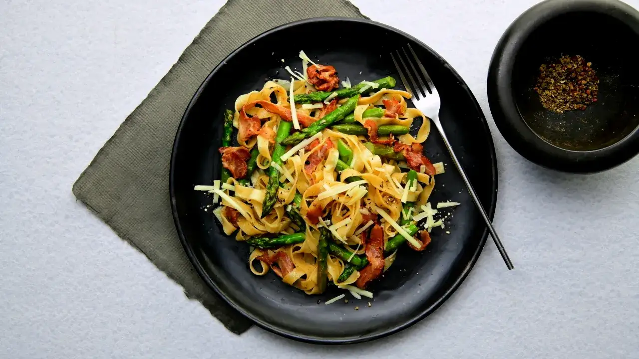 Pyszne spaghetti ze szparagami i chrupiącym boczkiem, posypane parmezanem. Obok moździerz z pieprzem.