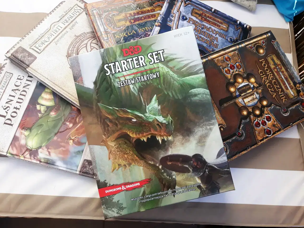 Zestaw startowy Dungeons & Dragons z zielonym smokiem i rycerzem. Obok inne podręczniki do gry.