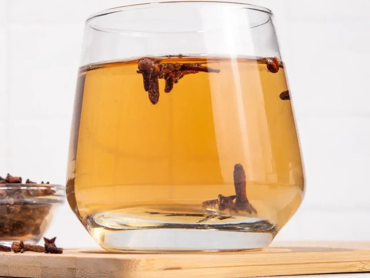 Szklanka z bursztynowym napojem, w którym pływają całe goździki. Obok miseczka z suszonymi goździkami.