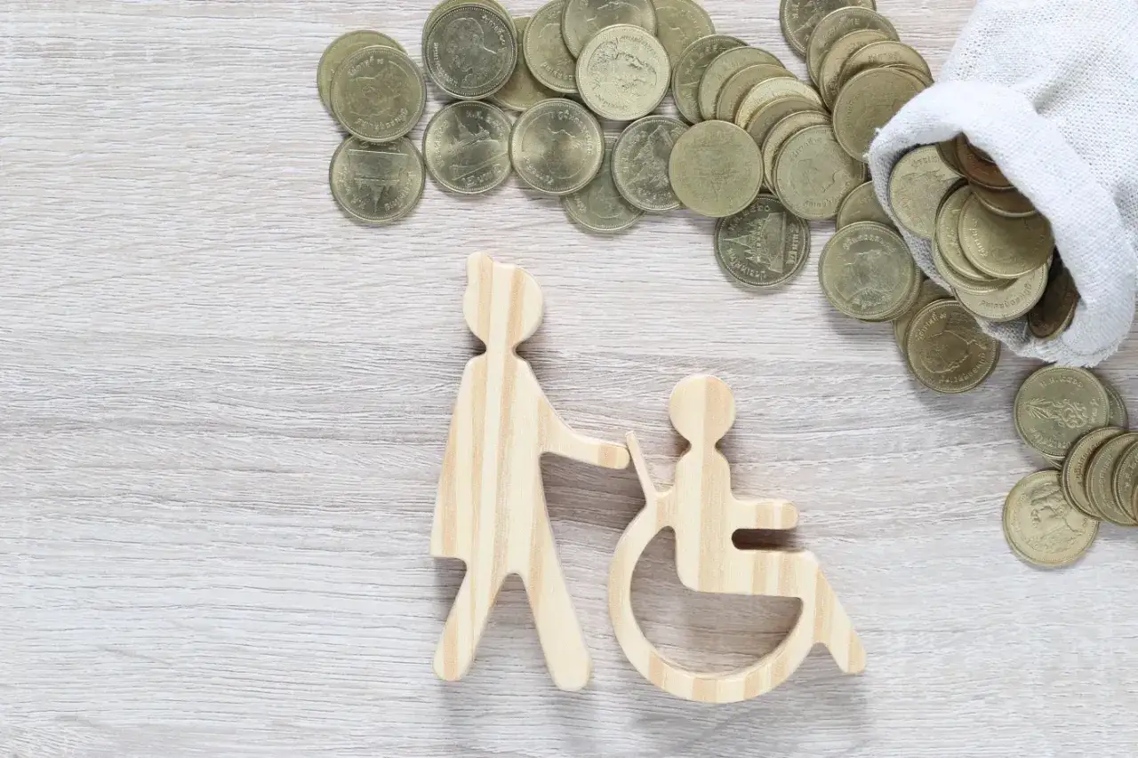 Drewniane figurki symbolizujące pomoc osobie na wózku inwalidzkim i stos monet. Finansowanie bezpłatnych wyjazdów dla osób niepełnosprawnych.