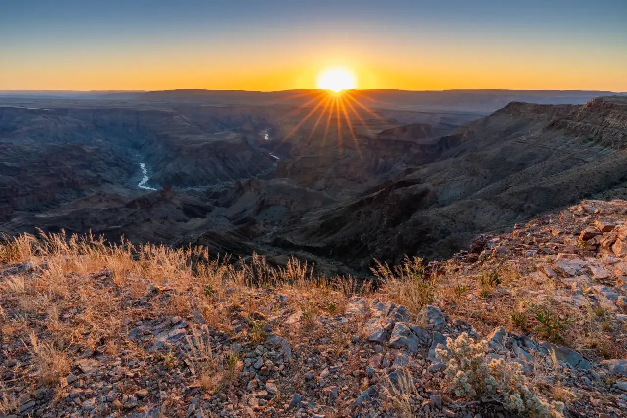 Zach&oacute;d słońca nad kanionem rzeki Fish. W dole wije się rzeka, a słońce rzuca długie cienie.