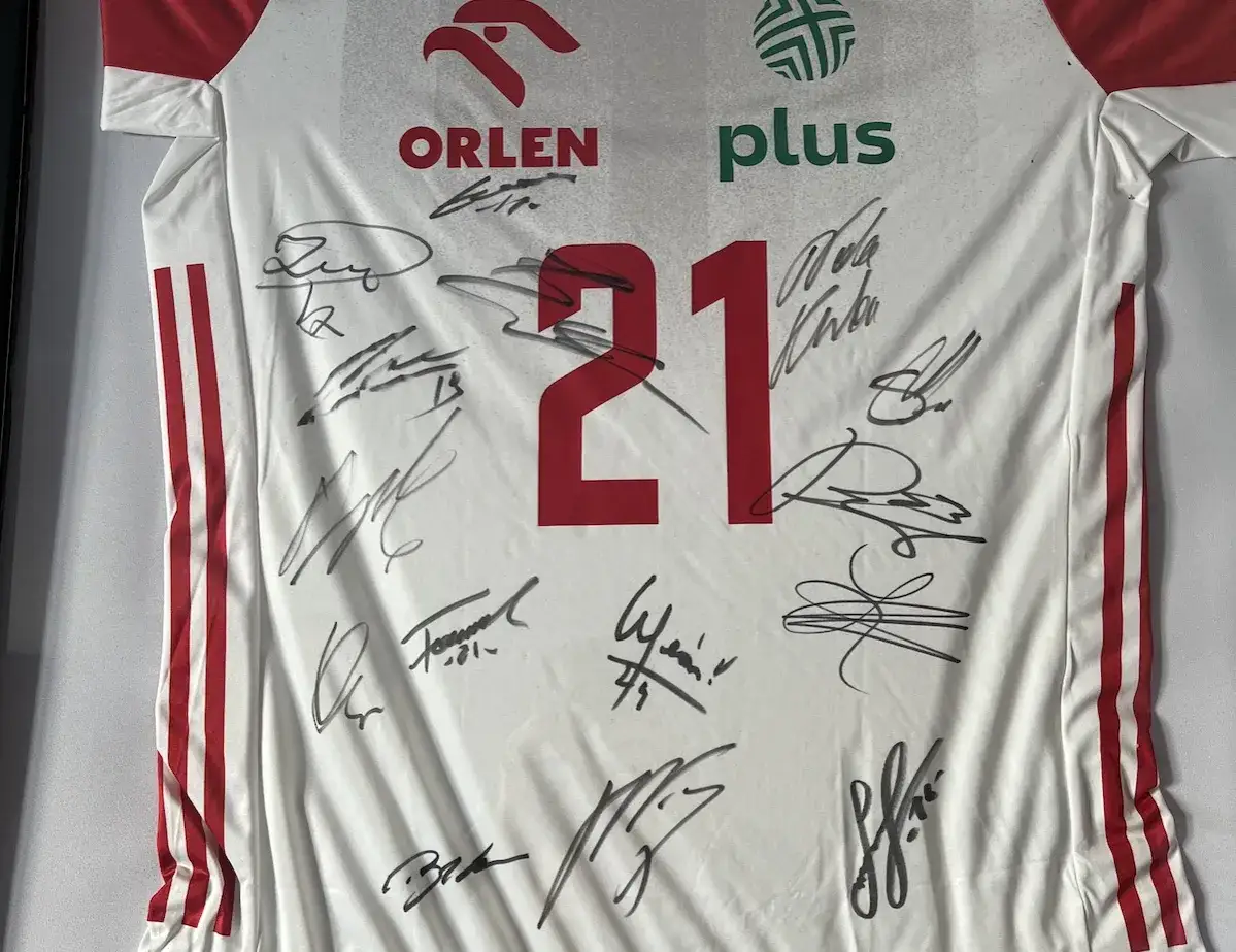 Biała koszulka siatkarska z czerwonymi pasami i numerem 21, pokryta licznymi autografami zawodnik&oacute;w.