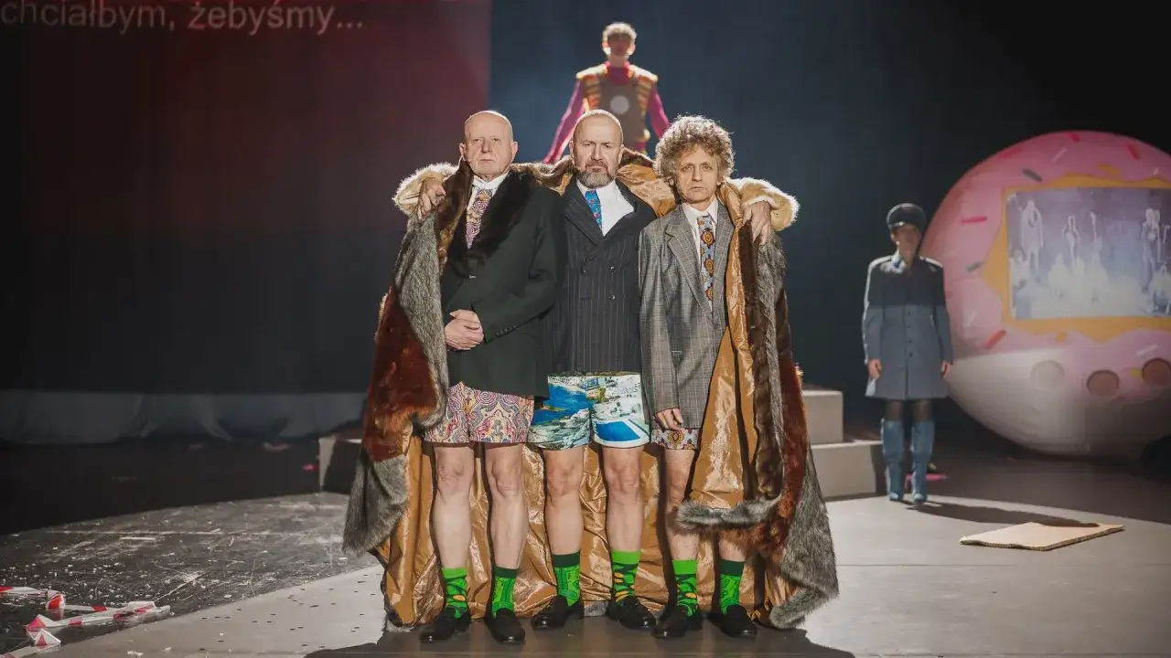 Trzech mężczyzn w futrach i kolorowych szortach, na scenie, podczas sztuki teatralnej po angielsku.