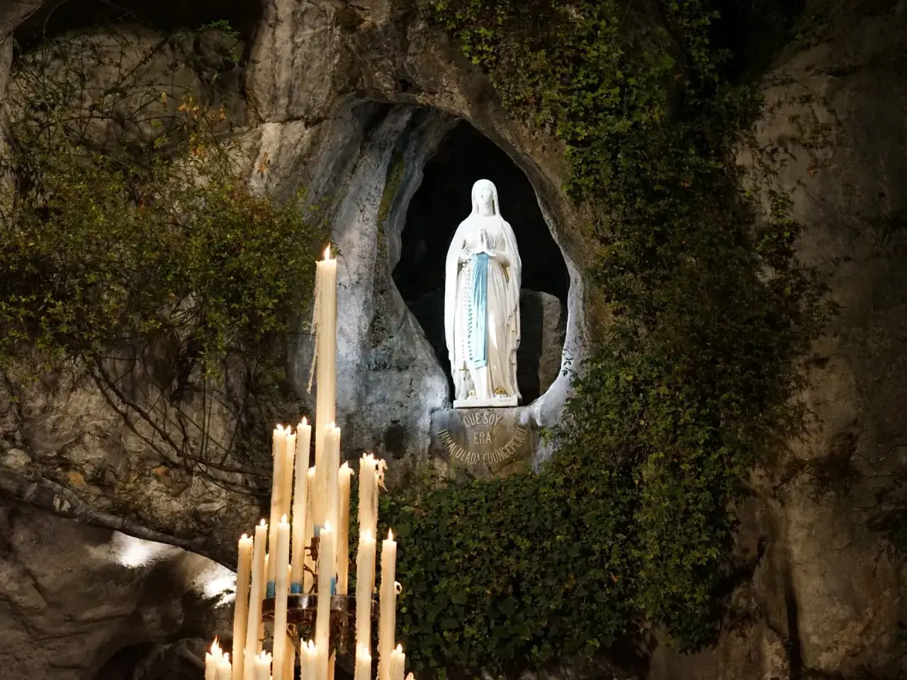 Figurka Matki Boskiej w grocie w Lourdes, otoczona płonącymi świecami. Jak znaleźć ciszę w Lourdes, mimo skutków komercjalizacji miejsc świętych?
