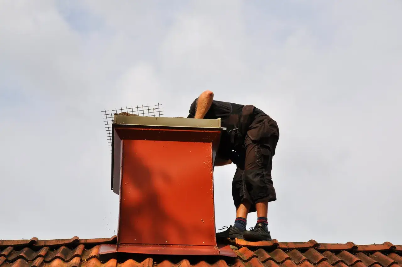 Pracownik na dachu uszczelnia komin z cegły, zabezpieczając go siatką.