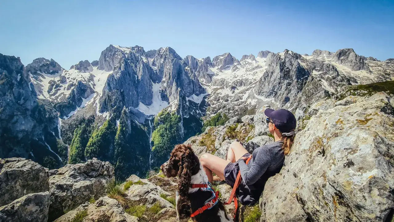 Kobieta z psem odpoczywa na skale, podziwiając majestatyczne, ośnieżone **g&oacute;ry przeklęte**.