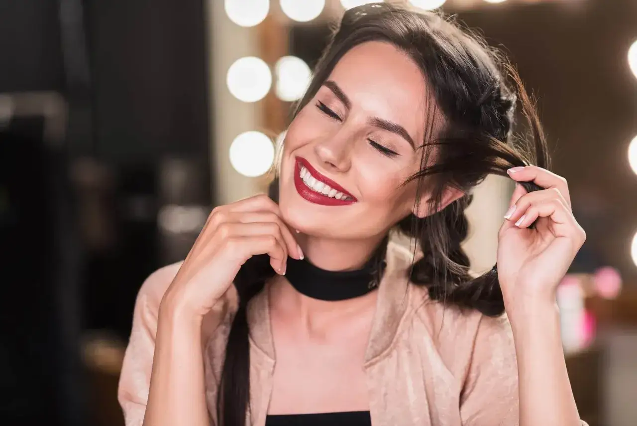 Uśmiechnięta dziewczyna z warkoczami, bawiąca się włosami. Jej spojrzenie i gesty to subtelne oznaki flirtu.