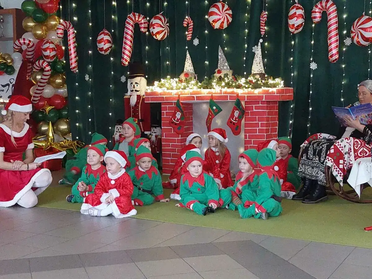 Wesołe **święta w przedszkolu**! Dzieci w strojach elf&oacute;w i Mikołaj&oacute;w słuchają bajki przy świątecznej scenografii.