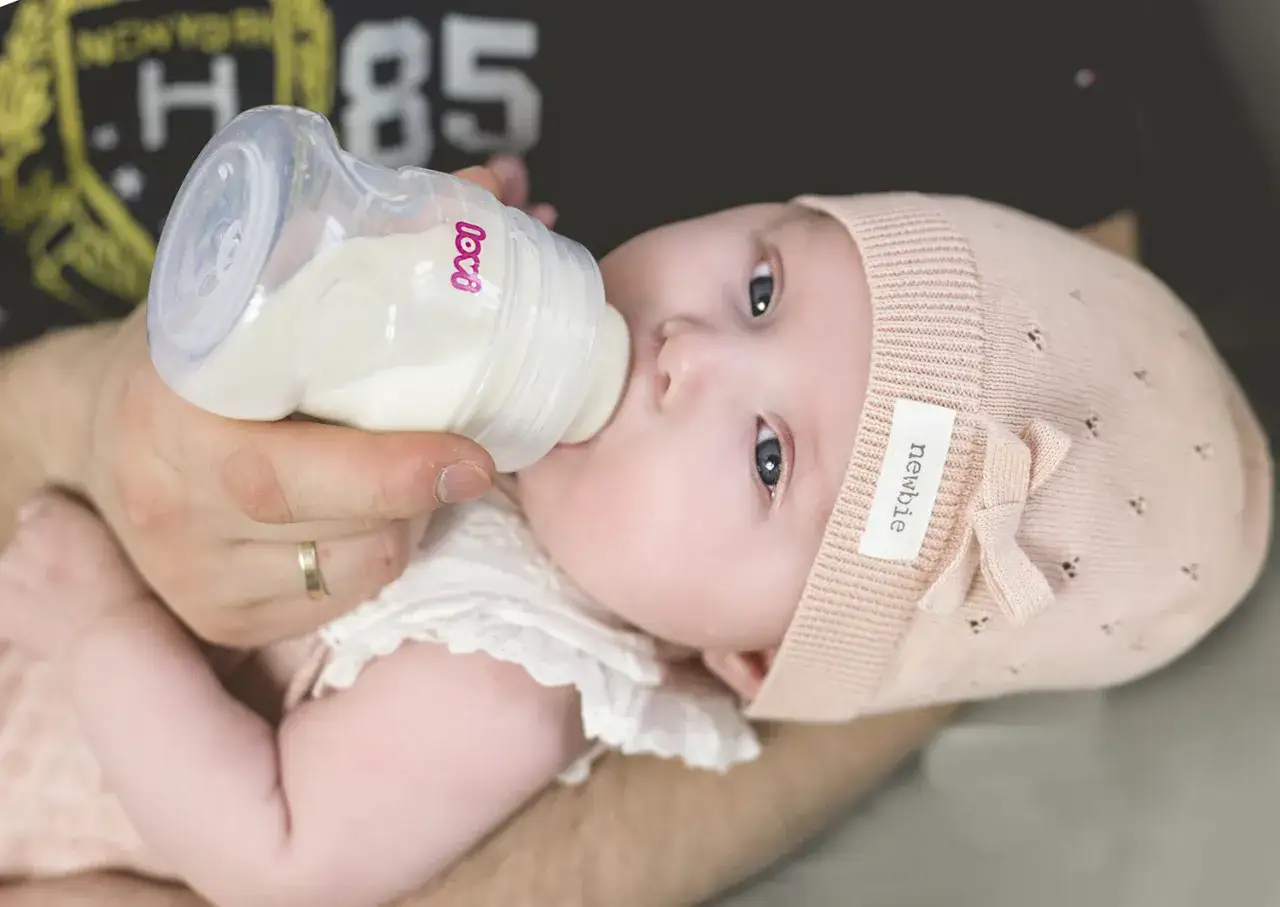 Dziecko w r&oacute;żowej czapeczce pije mleko z butelki. Karmienie mieszane to piękny moment bliskości.