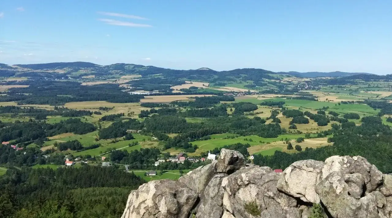 Widok z skał na malowniczą dolinę z lasami, polami i wioską. W oddali widać wzgórza, a na jednym z nich zarys dawnego zamku Bolczów.