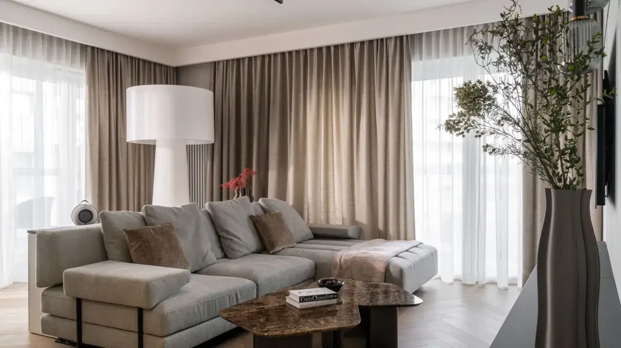 Eleganckie, beżowe zasłony do salonu tworzą przytulną atmosferę. Miękki narożnik i marmurowe stoliki dopełniają nowoczesny wystrój.