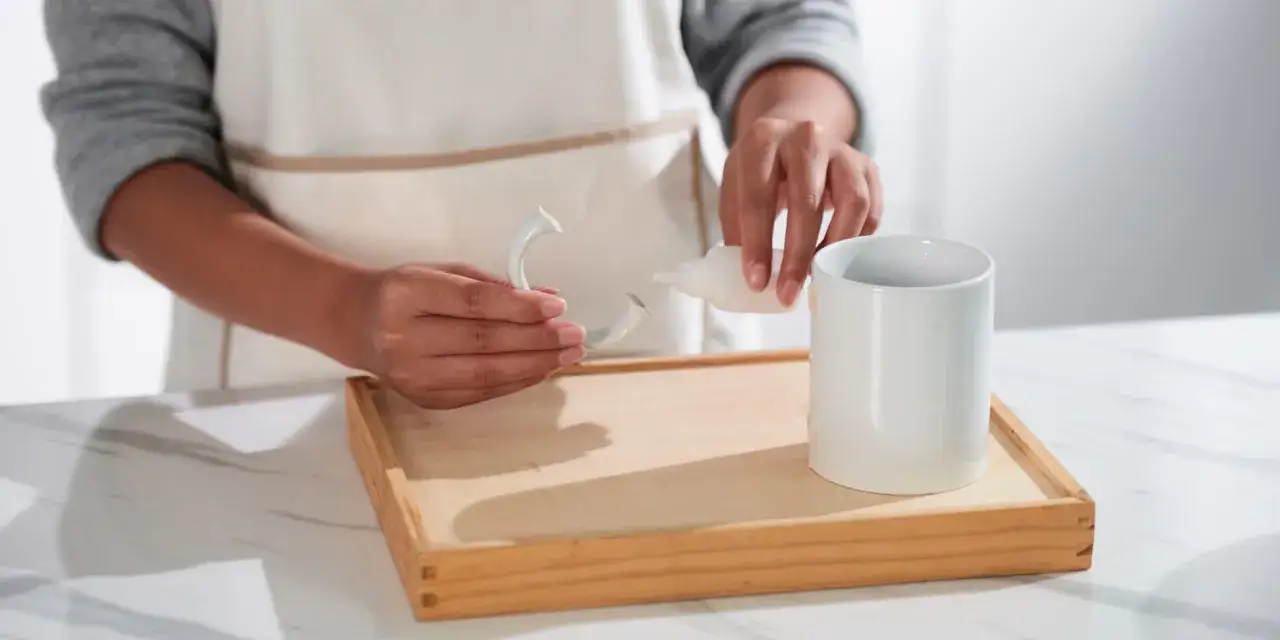 Osoba w fartuchu aplikuje klej na pęknięty uchwyt kubka ceramicznego, przygotowując się do jego naprawy.