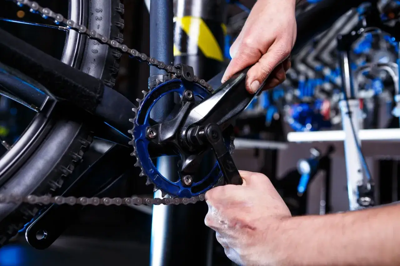Ręce majstrują przy rowerze, próbując zdjąć korbę bez ściągacza. Widoczny mechanizm korbowy i łańcuch.