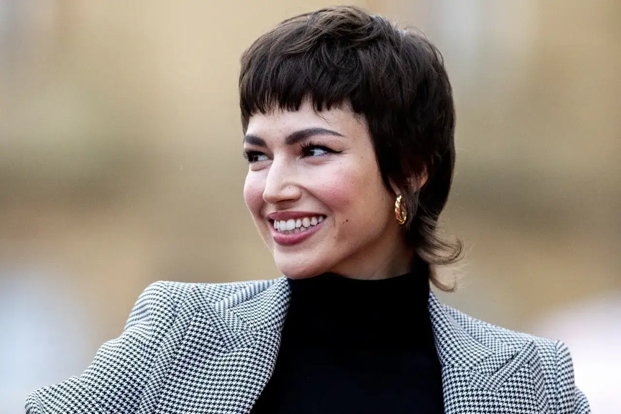 Uśmiechnięta kobieta z modną bixie fryzurą i złotymi kolczykami w kształcie k&oacute;łek, ubrana w czarny golf i dwukolorową marynarkę.