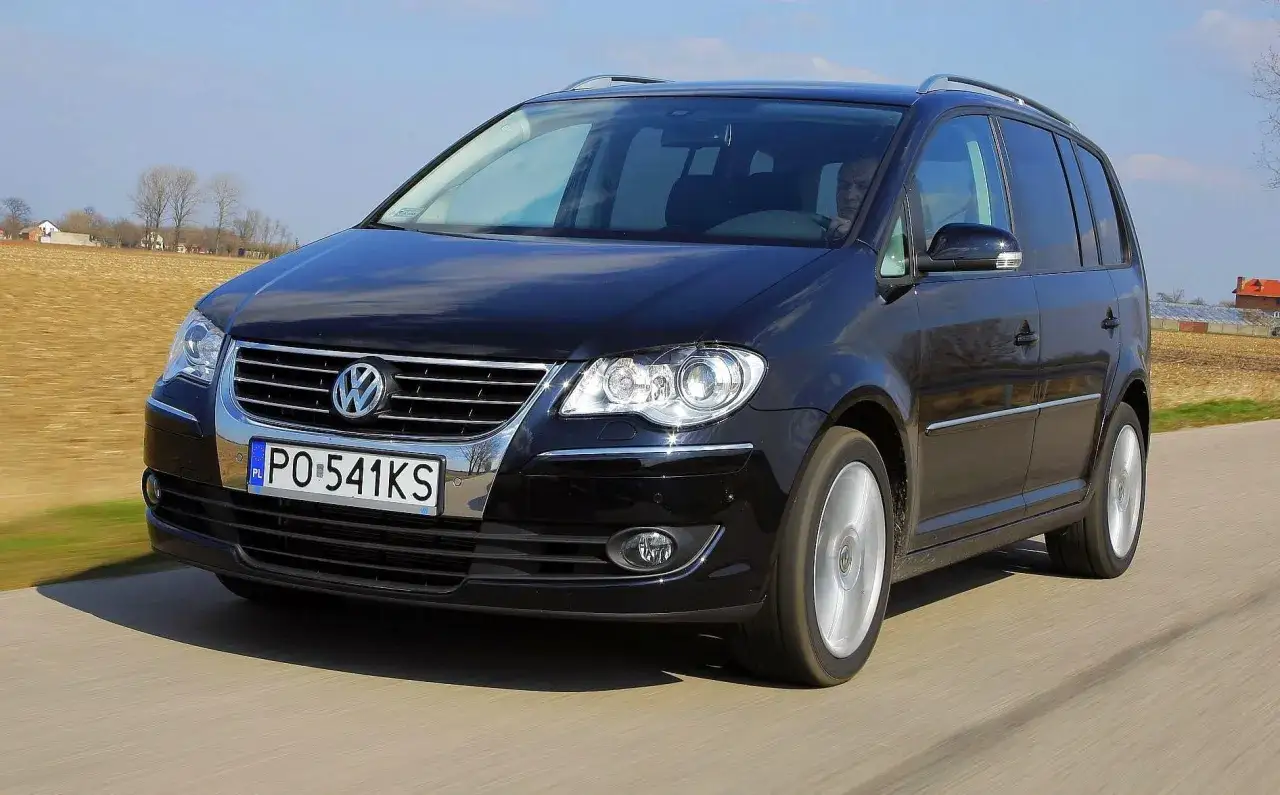 Czarny Volkswagen Touran jedzie po drodze, za nim pola i domy.