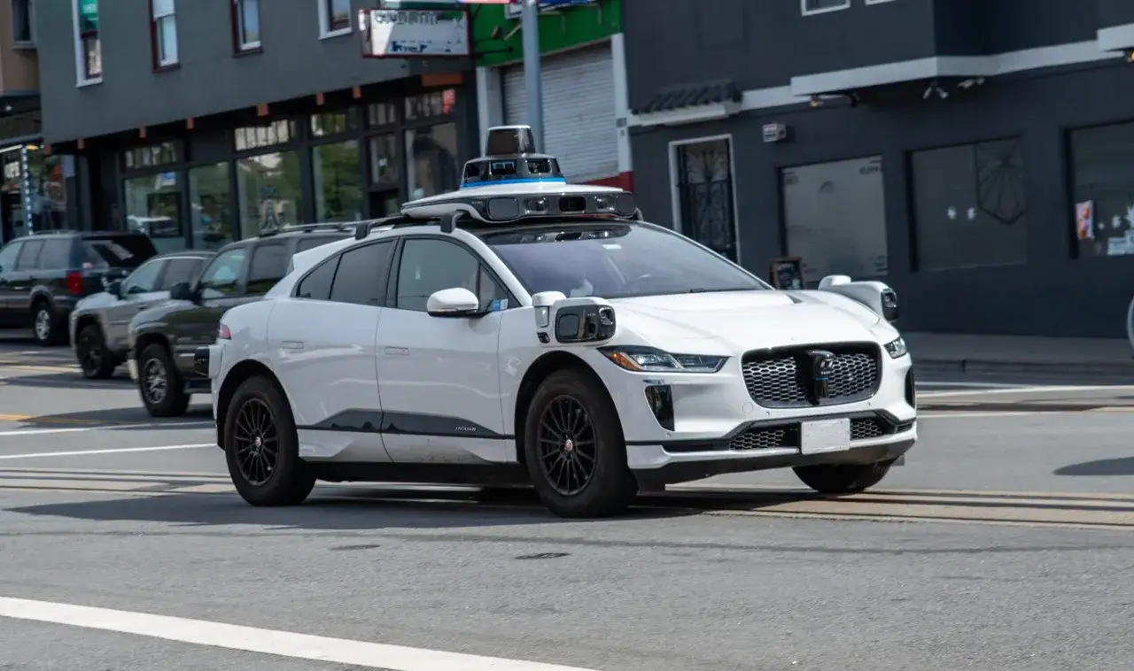 Biały Jaguar z czujnikami na dachu, jeden z autonomicznych pojazdów, jedzie ulicą w mieście.