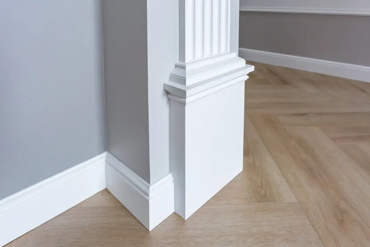 Biały pilaster, co to jest? To ozdobny element architektoniczny, który dodaje elegancji wnętrzu. Widoczny na zdjęciu przy szarej ścianie i drewnianej podłodze.
