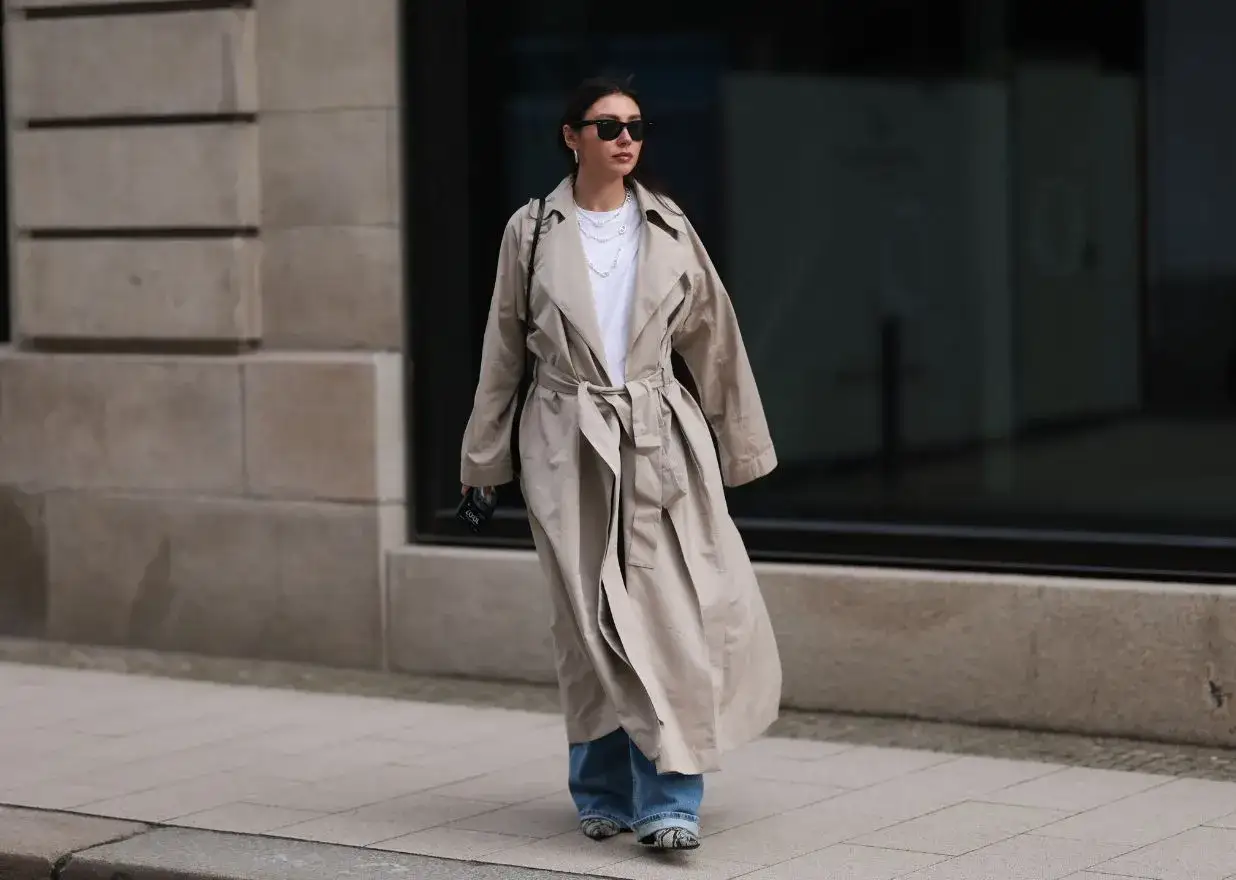 Kobieta w luźnych jeansach i białej koszulce prezentuje stylowy, jasny płaszcz. Jej stylizacje dopełniają okulary i naszyjniki.