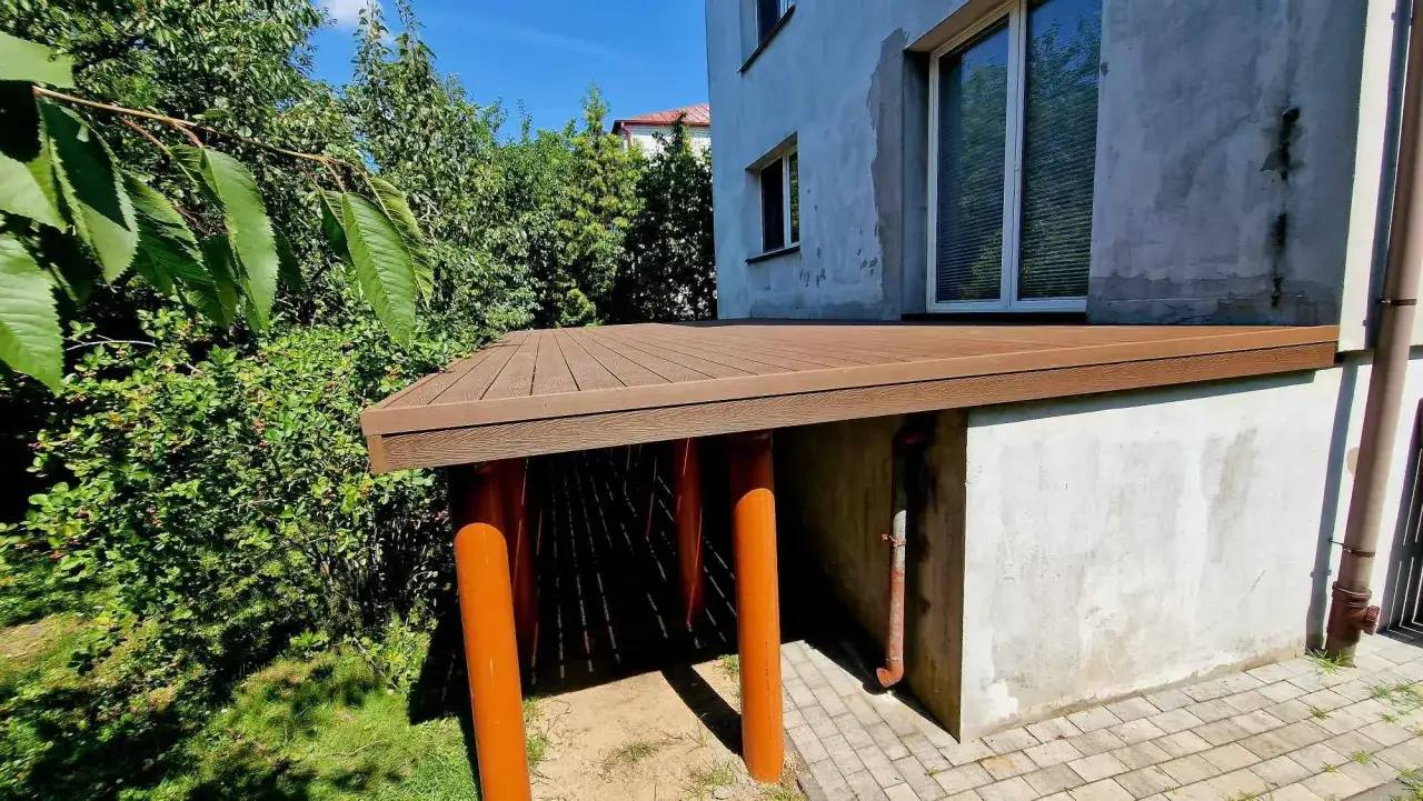 Nowy, wysoki taras na słupach, wykonany z brązowego kompozytu, stanowi idealne miejsce do wypoczynku w otoczeniu zieleni.