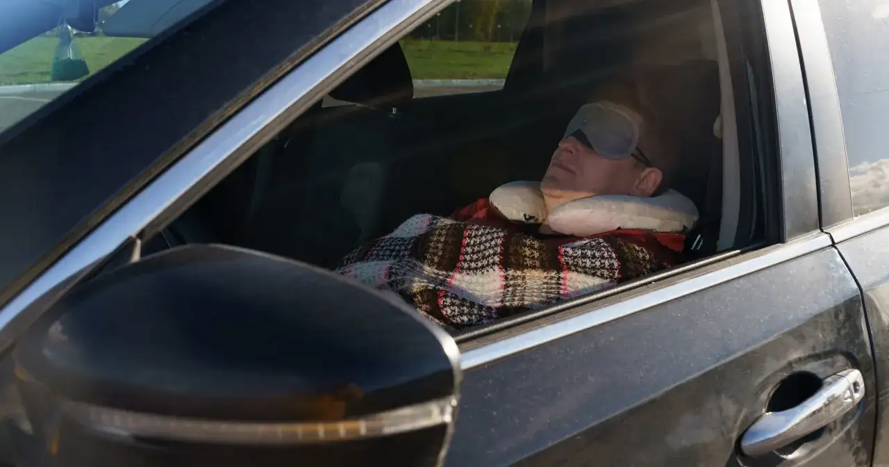 Mężczyzna śpi w samochodzie, otulony kocem i z poduszką pod szyją. To jego spos&oacute;b na spanie w samochodzie w Europie podczas podr&oacute;ży.