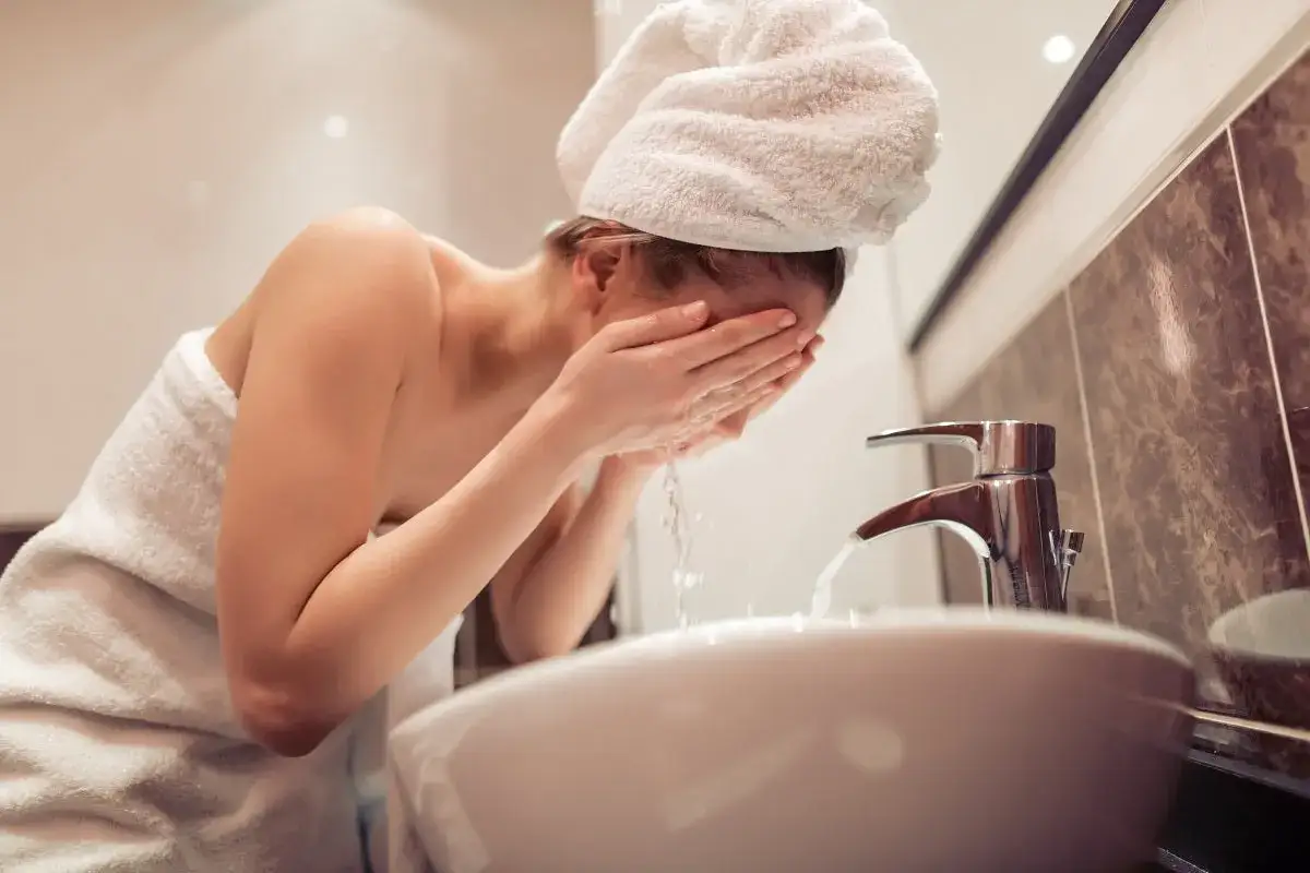 Kobieta w ręczniku na głowie i ciele, pochylona nad umywalką, wykonuje dwuetapowe oczyszczanie twarzy, spłukując wodą twarz.