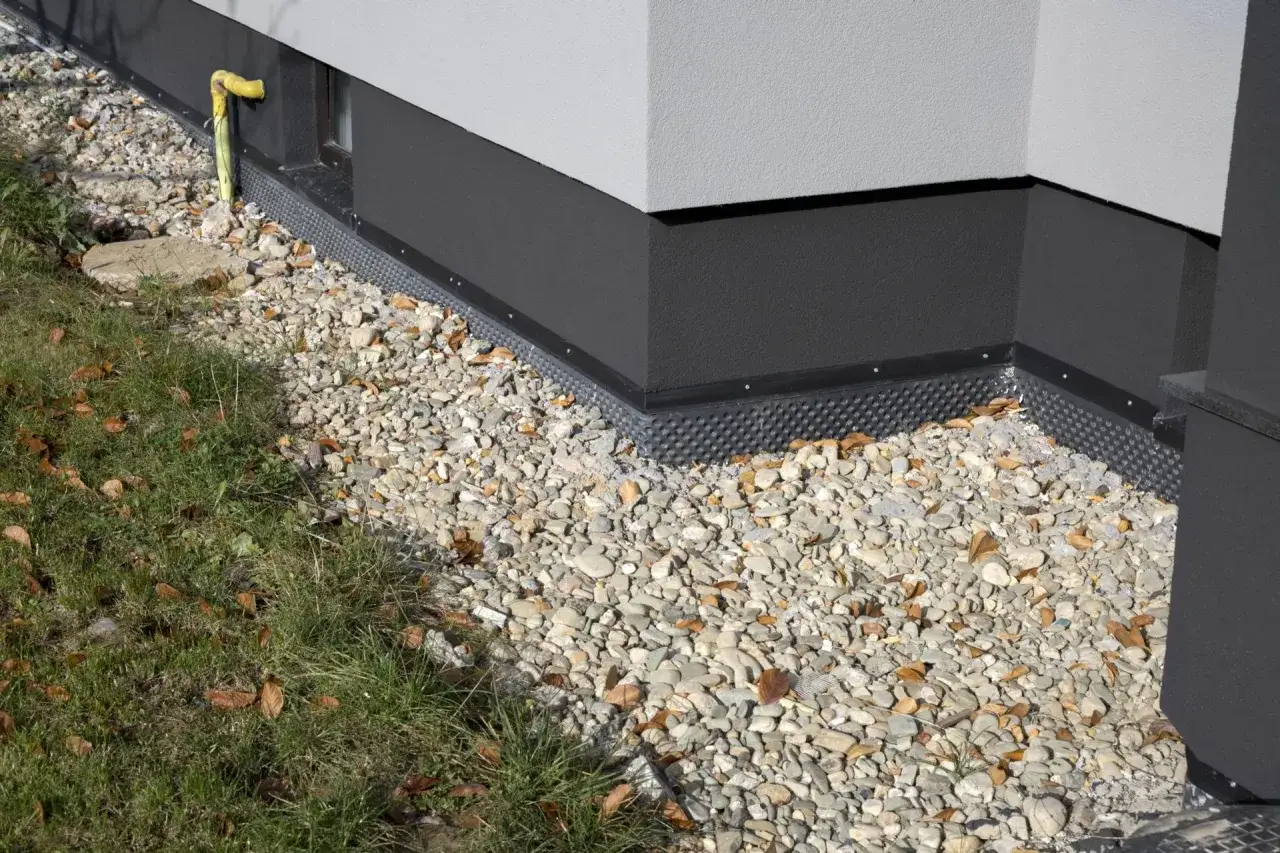 Folia kubełkowa na fundament chroni ścianę przed wilgocią. Widać ją pod warstwą kamieni i przy trawniku.