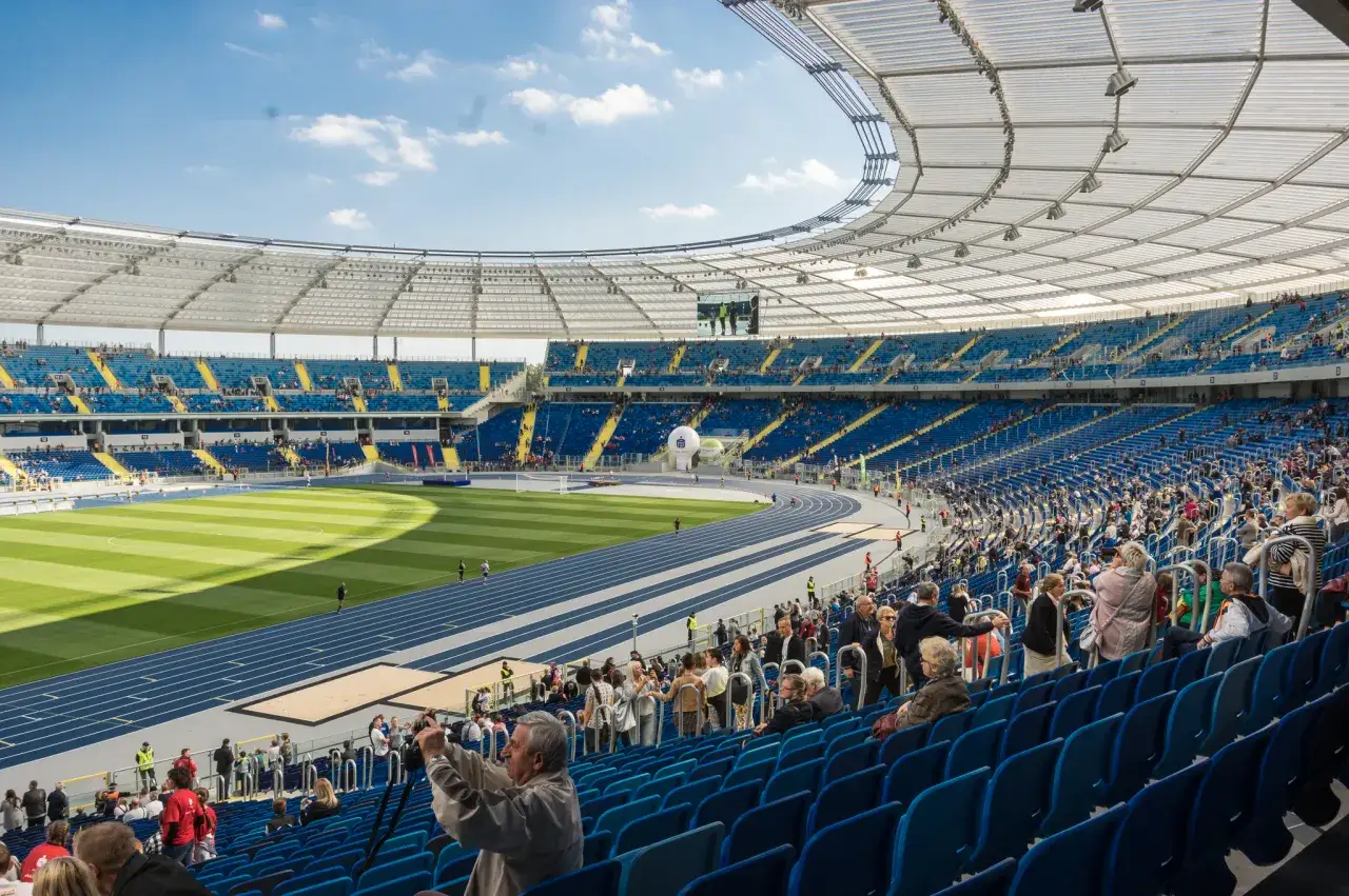 Widok na trybuny i bieżnię Stadionu Śląskiego w Chorzowie. Słońce oświetla zieloną murawę i niebieskie siedzenia.