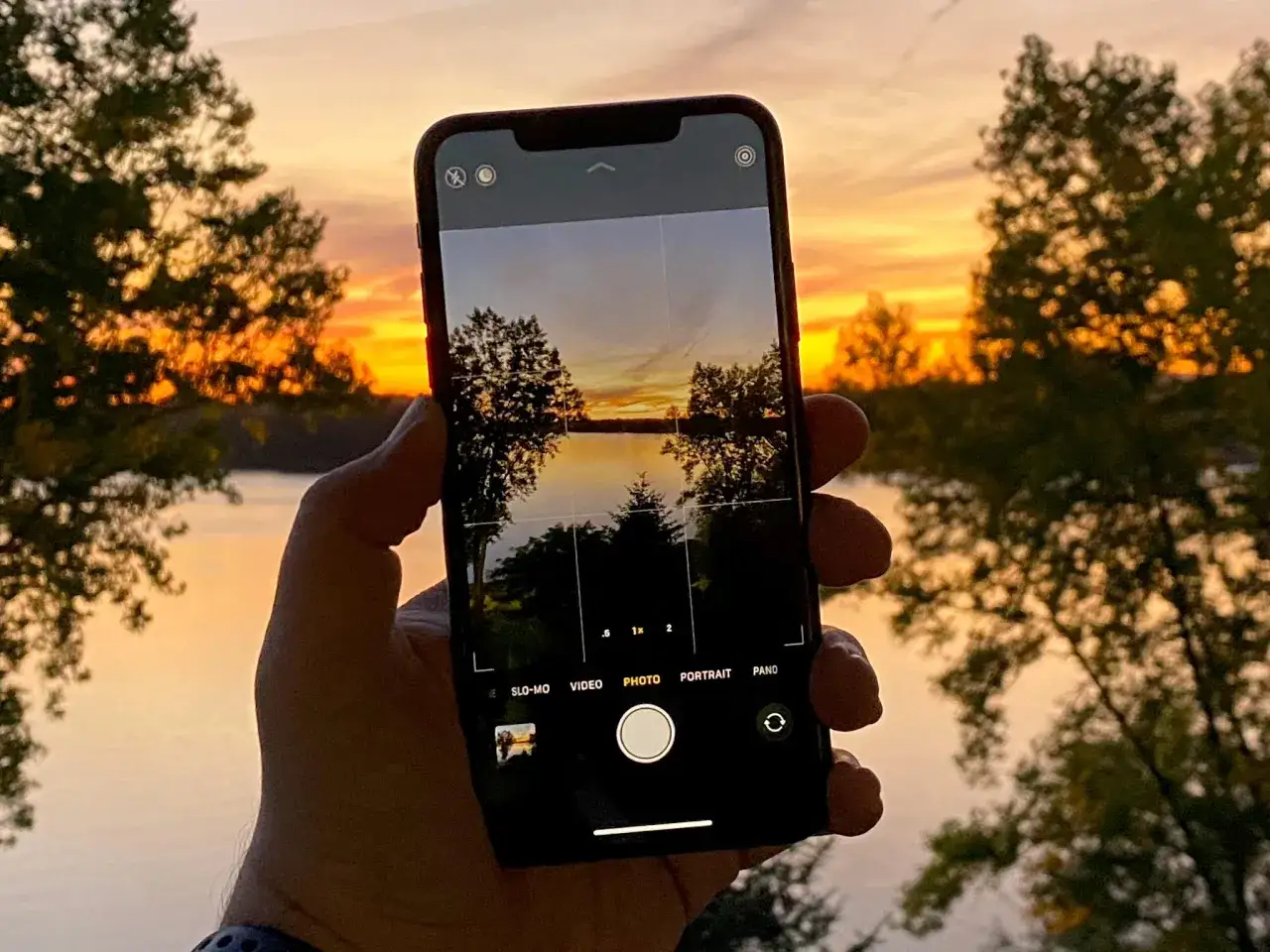 Dłoń trzyma iPhone 11, aparat uchwycił zachód słońca nad jeziorem.