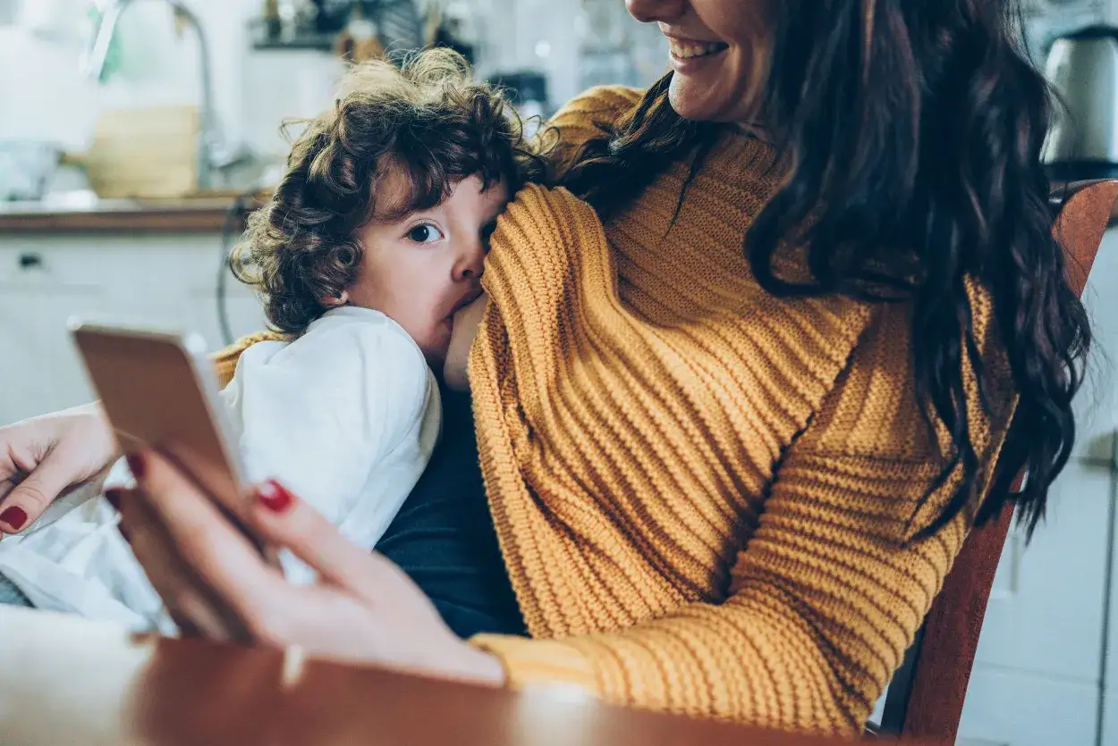 Mama z telefonem w ręku karmi piersią dziecko z kręconymi włosami. To moment, gdy szuka porad, jak odstawić dziecko od piersi.