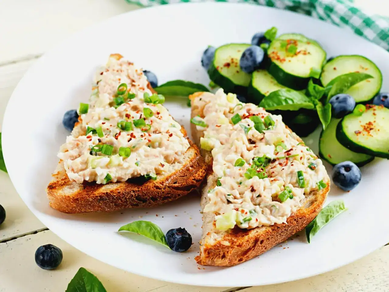 Dwa tosty z chleba pełnoziarnistego z pyszną pastą z tuńczyka, posypane szczypiorkiem i papryką. Obok plasterki ogórka i borówki.