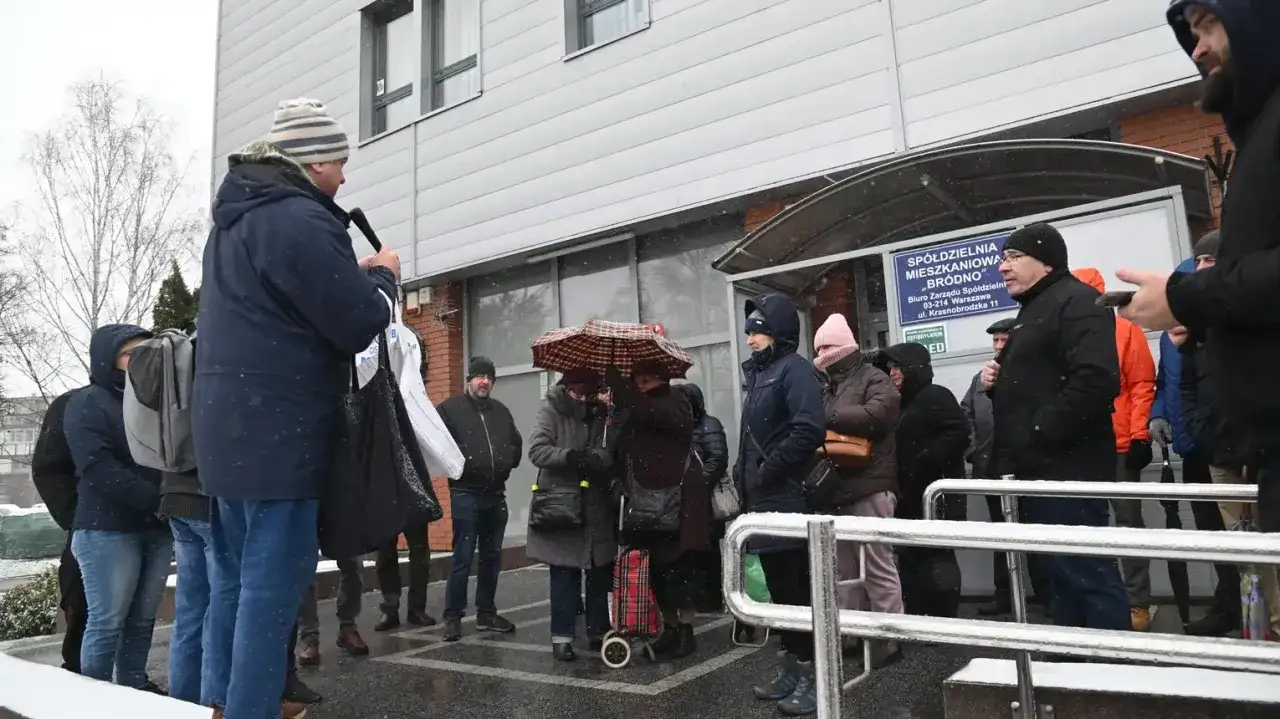 Ludzie stoją przed budynkiem spółdzielni mieszkaniowej. Dyskusja o tym, ile trwa kadencja prezesa spółdzielni mieszkaniowej, toczy się w zimowej aurze.