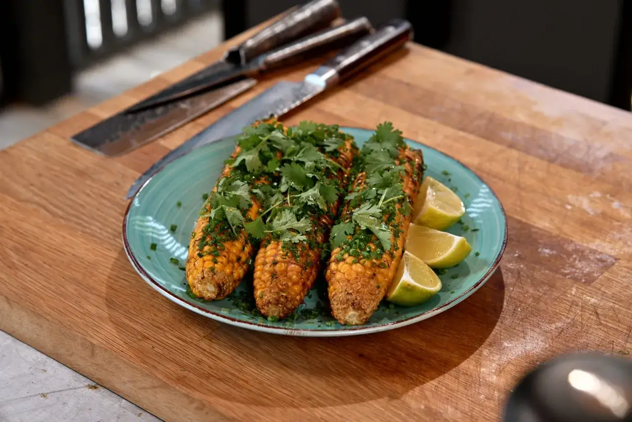 Trzy kolby kukurydzy z grilla, posypane szczypiorkiem i kolendrą, podane z ćwiartkami limonki na turkusowym talerzu.