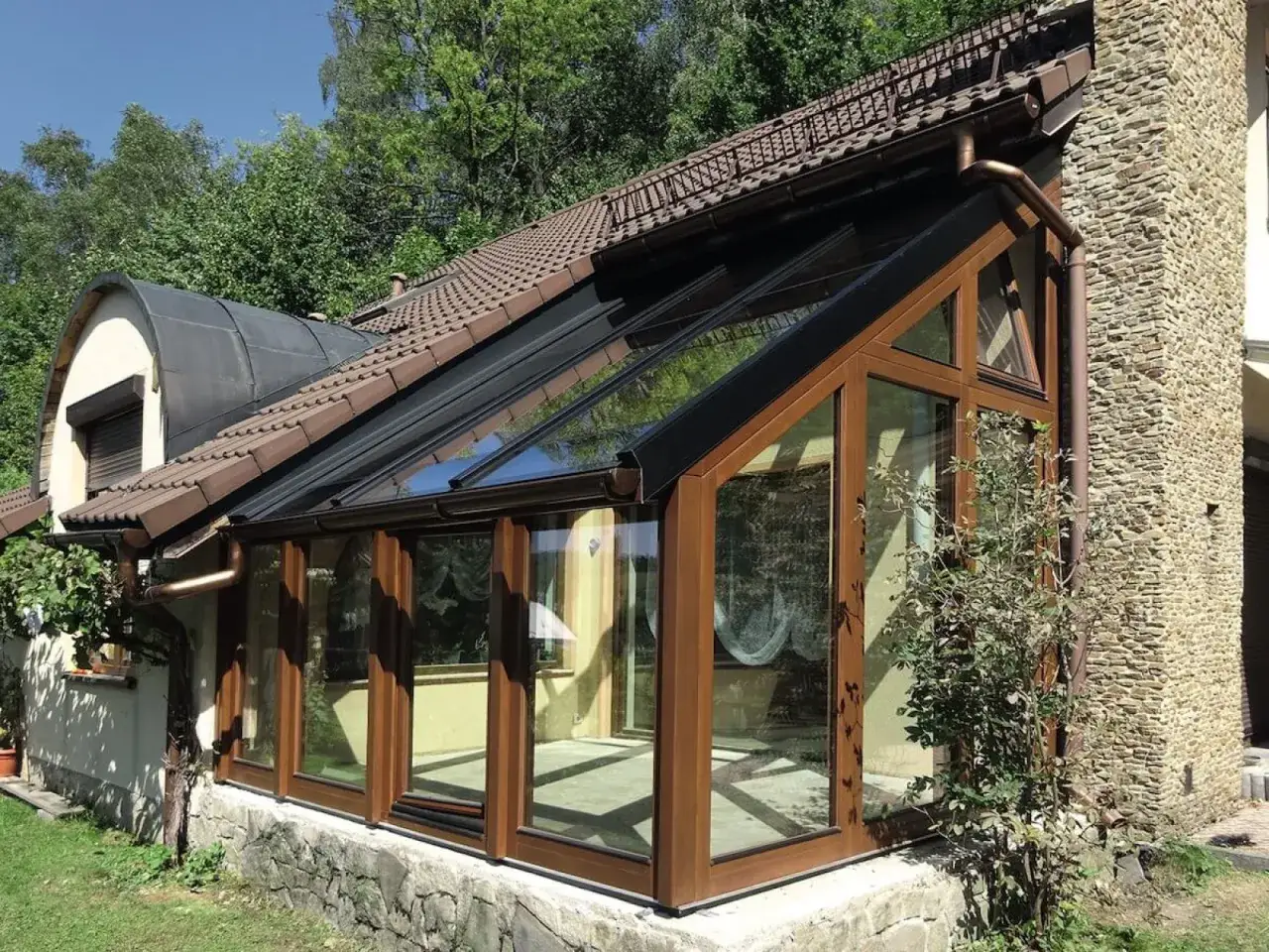 Nowoczesny ogród zimowy do samodzielnego montażu, z drewnianymi ramami i przeszklonym dachem, przylega do kamiennego domu.