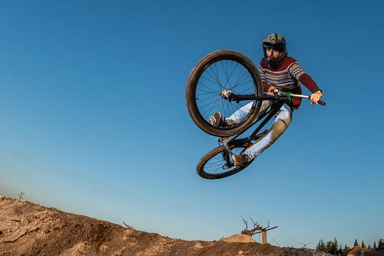 Kolarz w kasku wykonuje efektowne hopy rowerowe, unosząc się w powietrzu na tle błękitnego nieba.