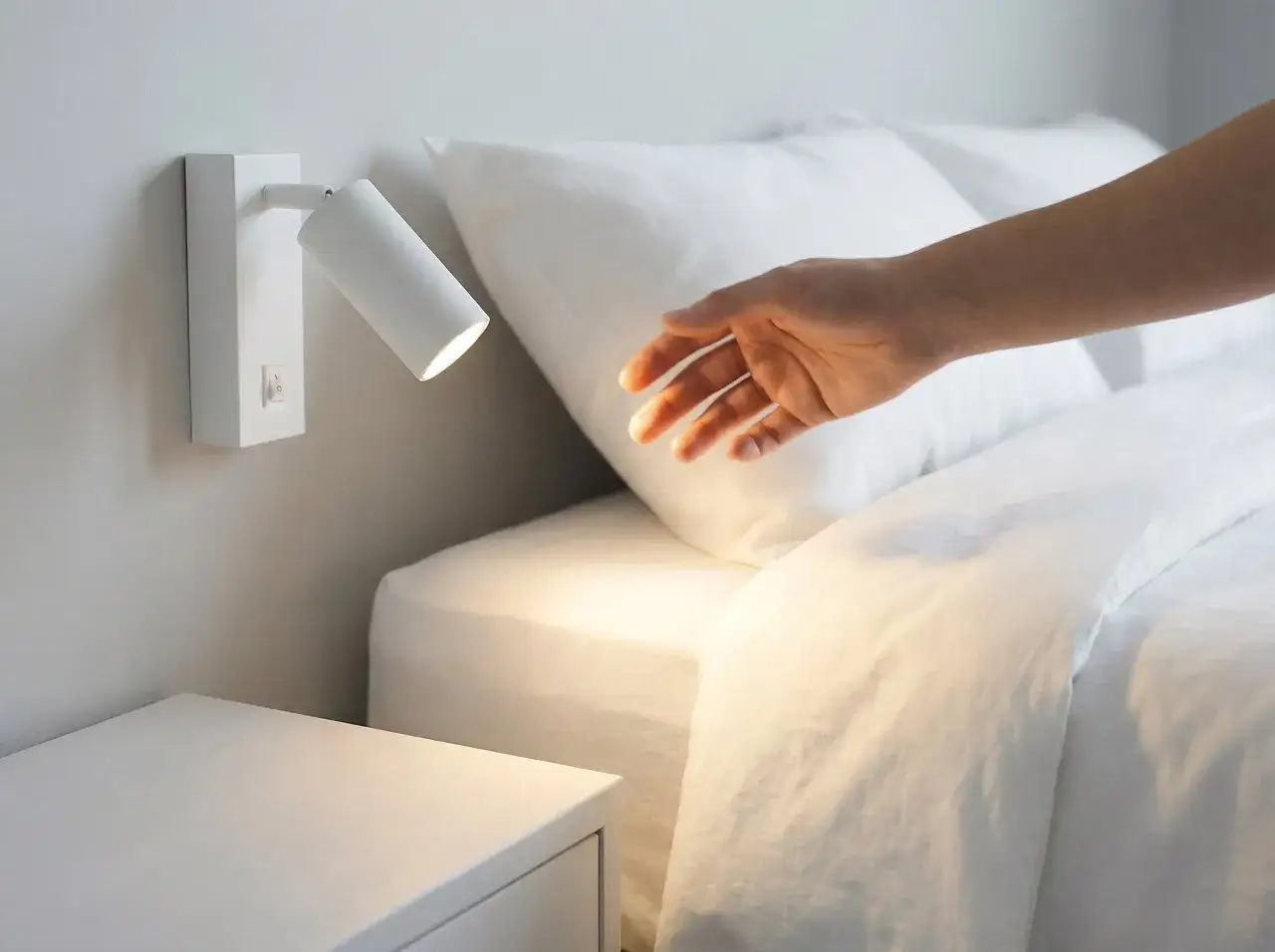 Białe kinkiety do sypialni nad łóżko, idealne do czytania. Dłoń sięga do włącznika, by stworzyć przytulną atmosferę.