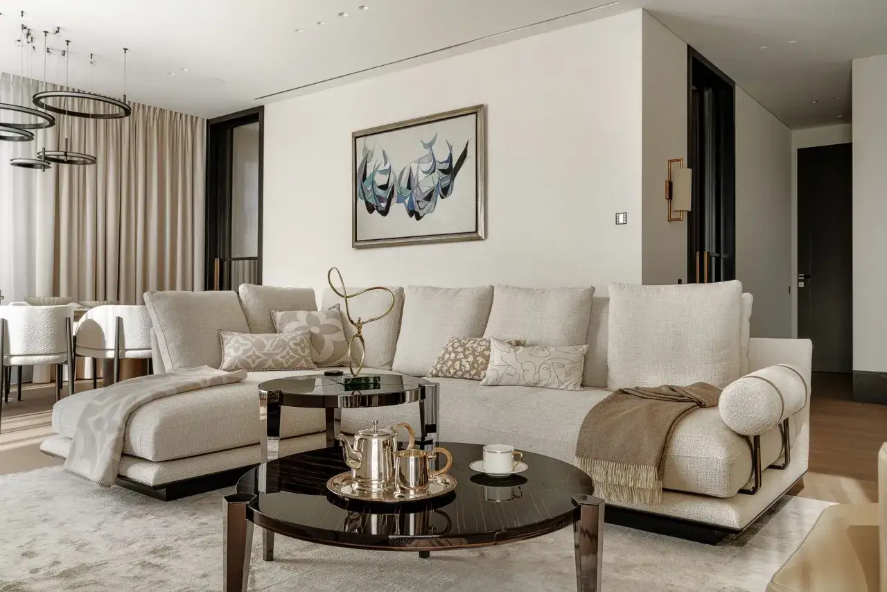 Przytulny salon w kolorze ecru. Duża, wygodna kanapa, stoliki kawowe z błyszczącym blatem, srebrny serwis do herbaty i obraz na ścianie tworzą elegancką przestrzeń.