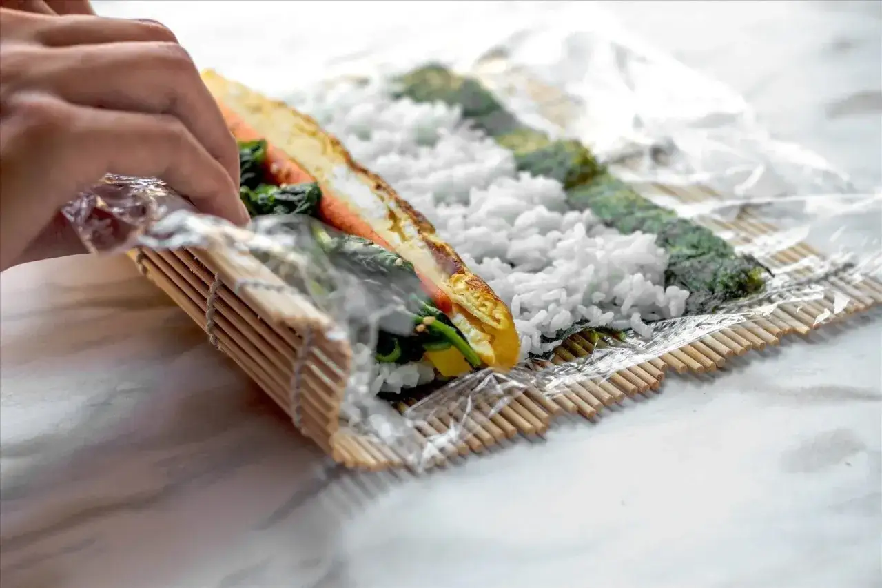 Ręce zwijają sushi na macie bambusowej. Widać ryż, nori i kolorowe nadzienie do sushi: omlet, szpinak i marchewkę.